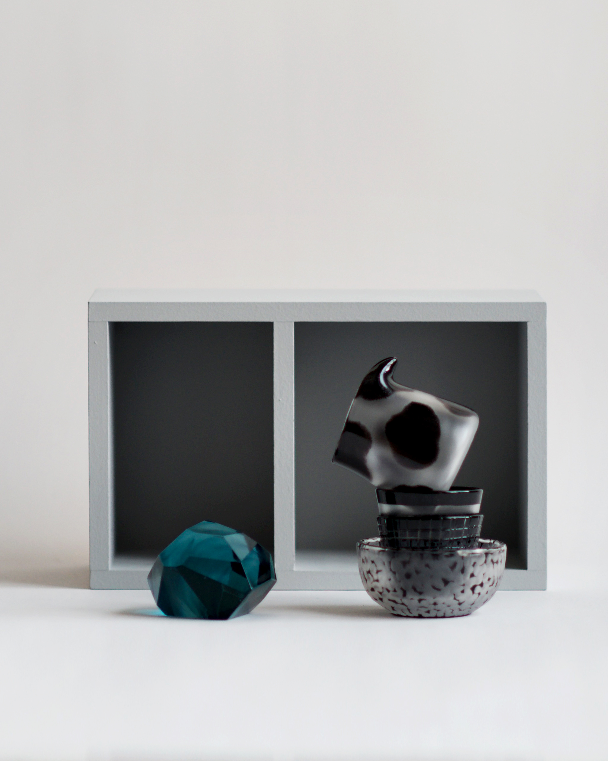 Composition by Factory Zoomer showcasing a light gray display shelf in the background and in the foreground a sapphire glass gem on the left with a staff of glass tableware on the right