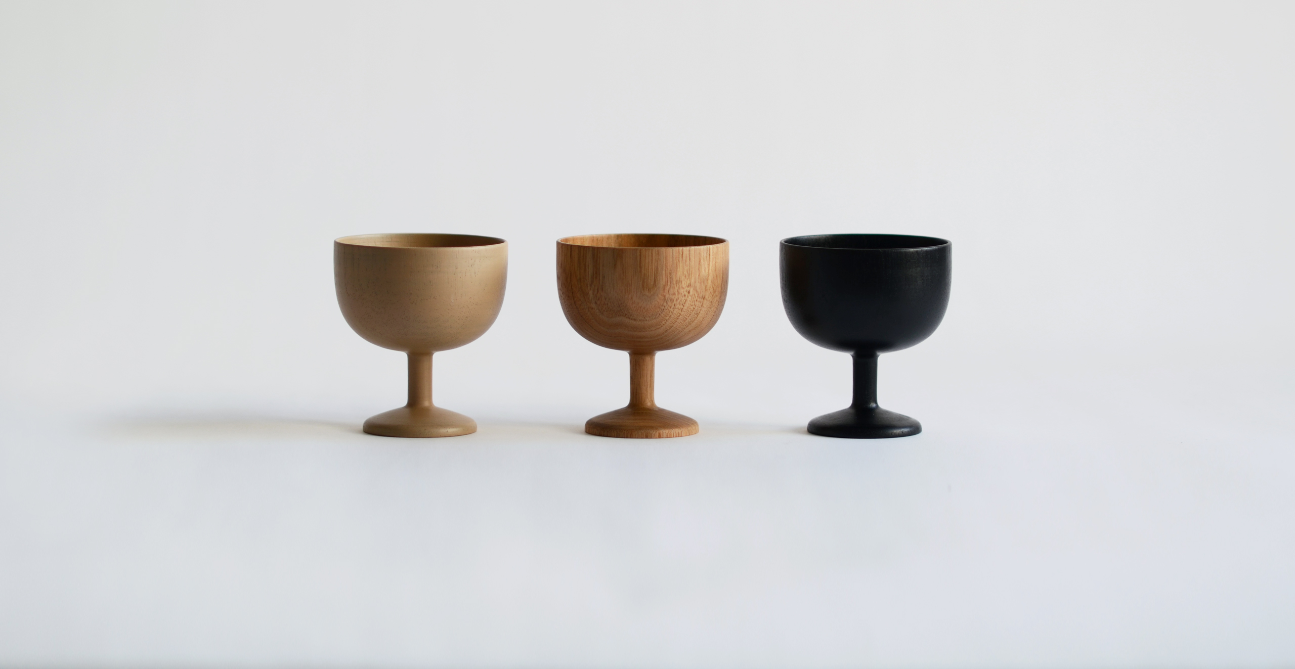 three wooden ice cream cups in a row against an offwhite background. 1 is urushi lacquered, one is wood and one is black