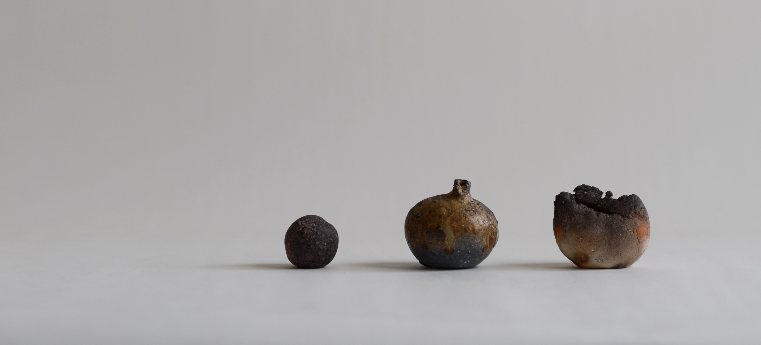 Three small wood fired pieces, astone vase, vase and textured shell by Keisuke Iwata in a row against a light gray backdround