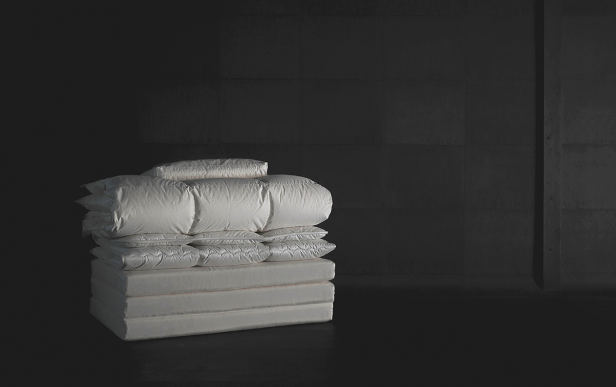 dark image of offwhite futon and fluffy down duvet setup by Iwata Nezen stacked and folded in a neat pile in a dark room